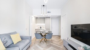 Un salon moderne avec un canapé gris, une table à manger avec des chaises, un téléviseur et, au fond, un coin cuisine.