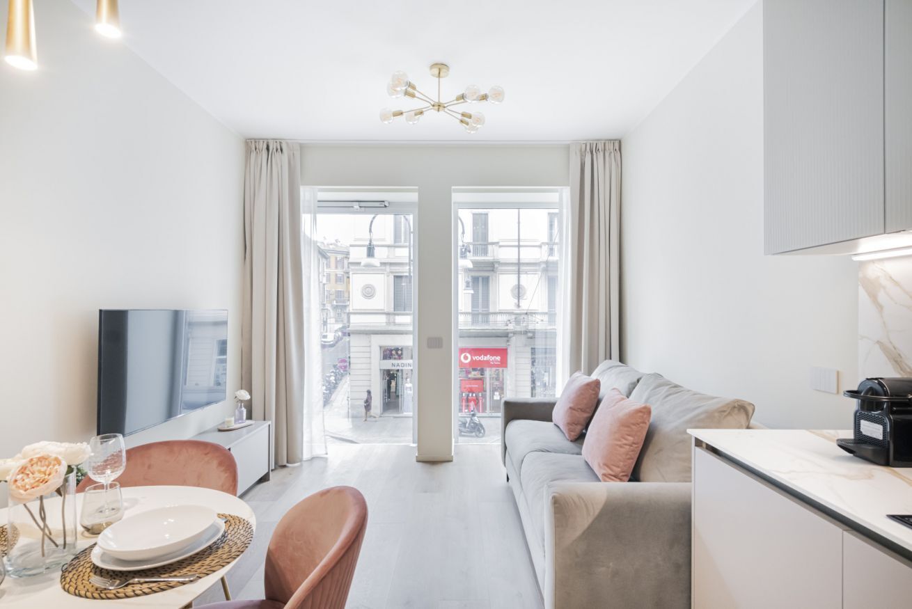 En moderne stue med sofa, spisebord og stole. Et fjernsyn står på et skab, og et stort vindue er dækket af gardiner.