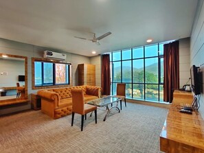 A spacious living room with a large window, a sofa, a glass coffee table, and a wooden desk. - Hotel Prashant International (Gandhidham)