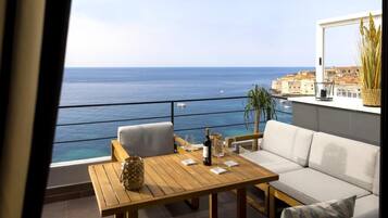 A balcony with a wooden table and two chairs, overlooking the sea and a coastal town.
