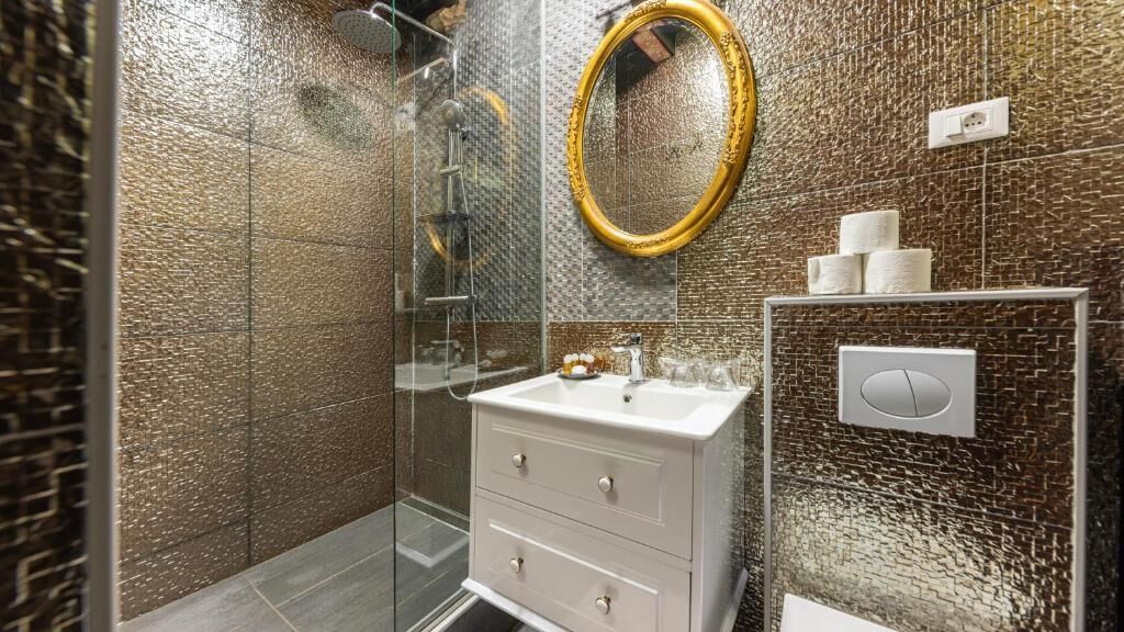 A bathroom with a glass shower enclosure, a white vanity with drawers, a gold-framed mirror, and a toilet with a paper holder.