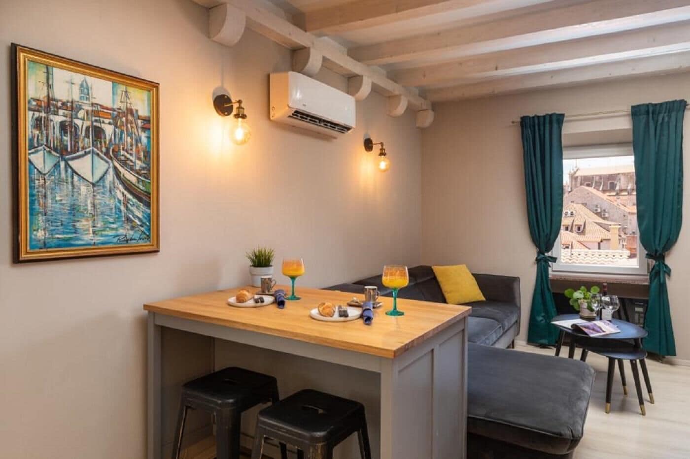 Un cuarto acogedor con una mesa de madera, dos taburetes negros, un sofá con un cojín amarillo, un cuadro en la pared y una ventana con cortinas.