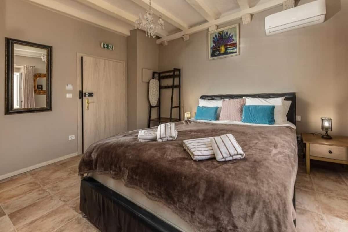 Un dormitorio con una cama grande, una mesita de noche con lámpara, una escalera y un cuadro en la pared.