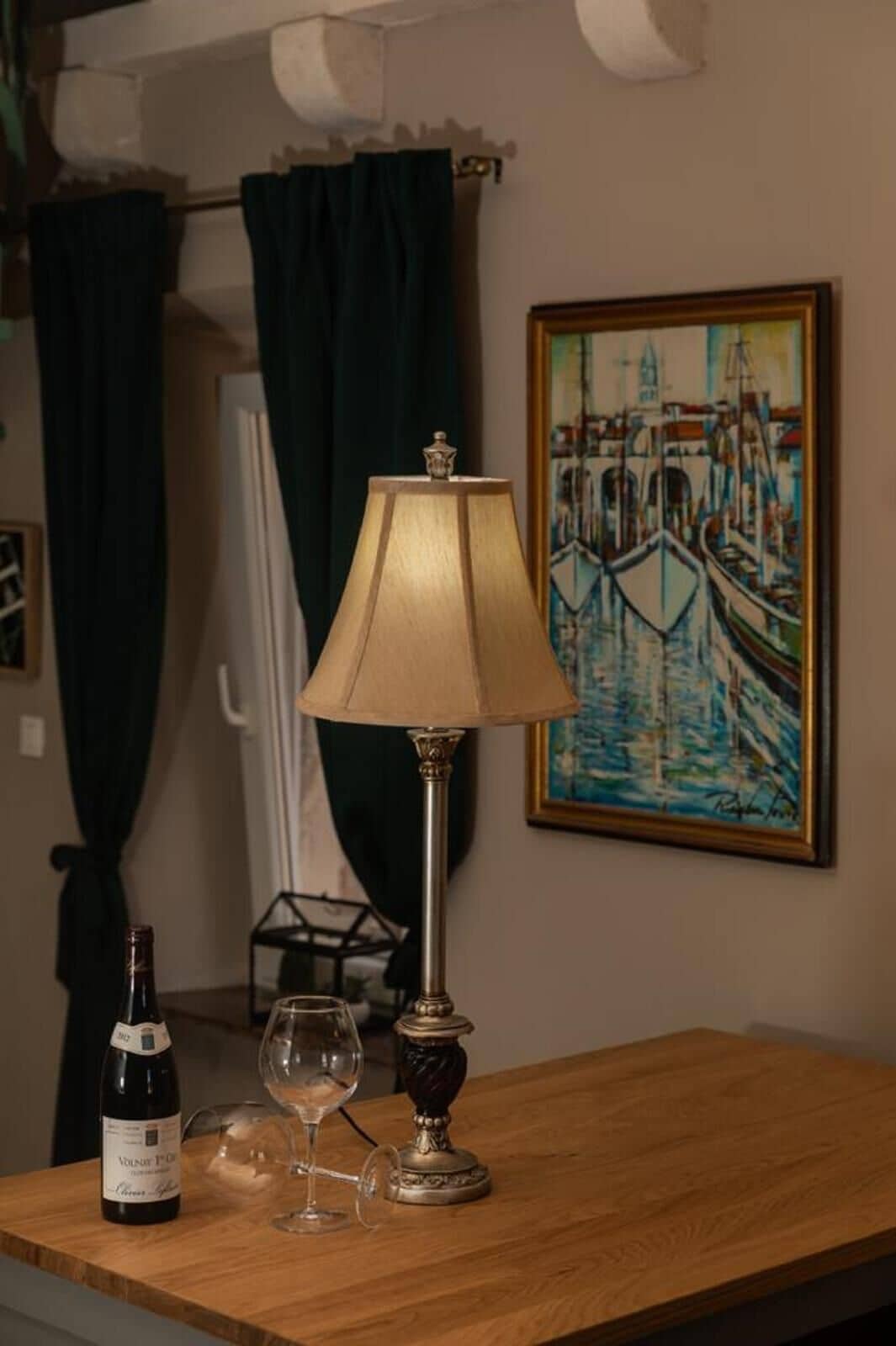 A wooden table with a lamp, a wine bottle, and a glass. A painting of a canal scene is on the wall.