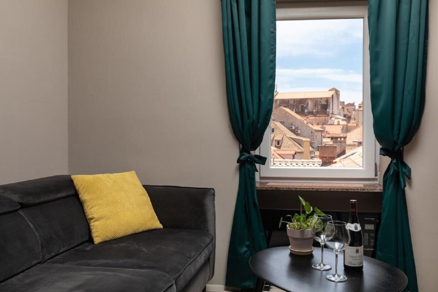 Une pièce avec un canapé noir, un coussin jaune, une fenêtre avec des rideaux verts, une plante en pot et une bouteille de vin posée sur une petite table.