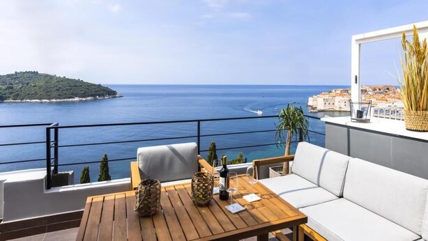 Un balcon avec une table en bois, un canapé blanc et une vue sur la mer et une île lointaine.