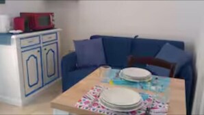 A small dining area with a table set for two, a blue sofa, and a white cabinet with a red microwave on top.