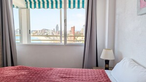 Habitació d'hotel amb un llit, una finestra amb cortines, una làmpada i vista a un paisatge urbà i una platja.
