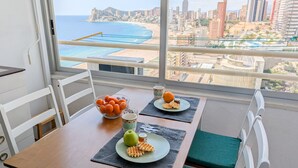 Zona de menjador amb una taula preparada per a dues persones, un bol d'albercocs, una poma verda, gofres i una tassa de cafè. L'habitació té vistes a una platja i un paisatge urbà a través de grans finestres.