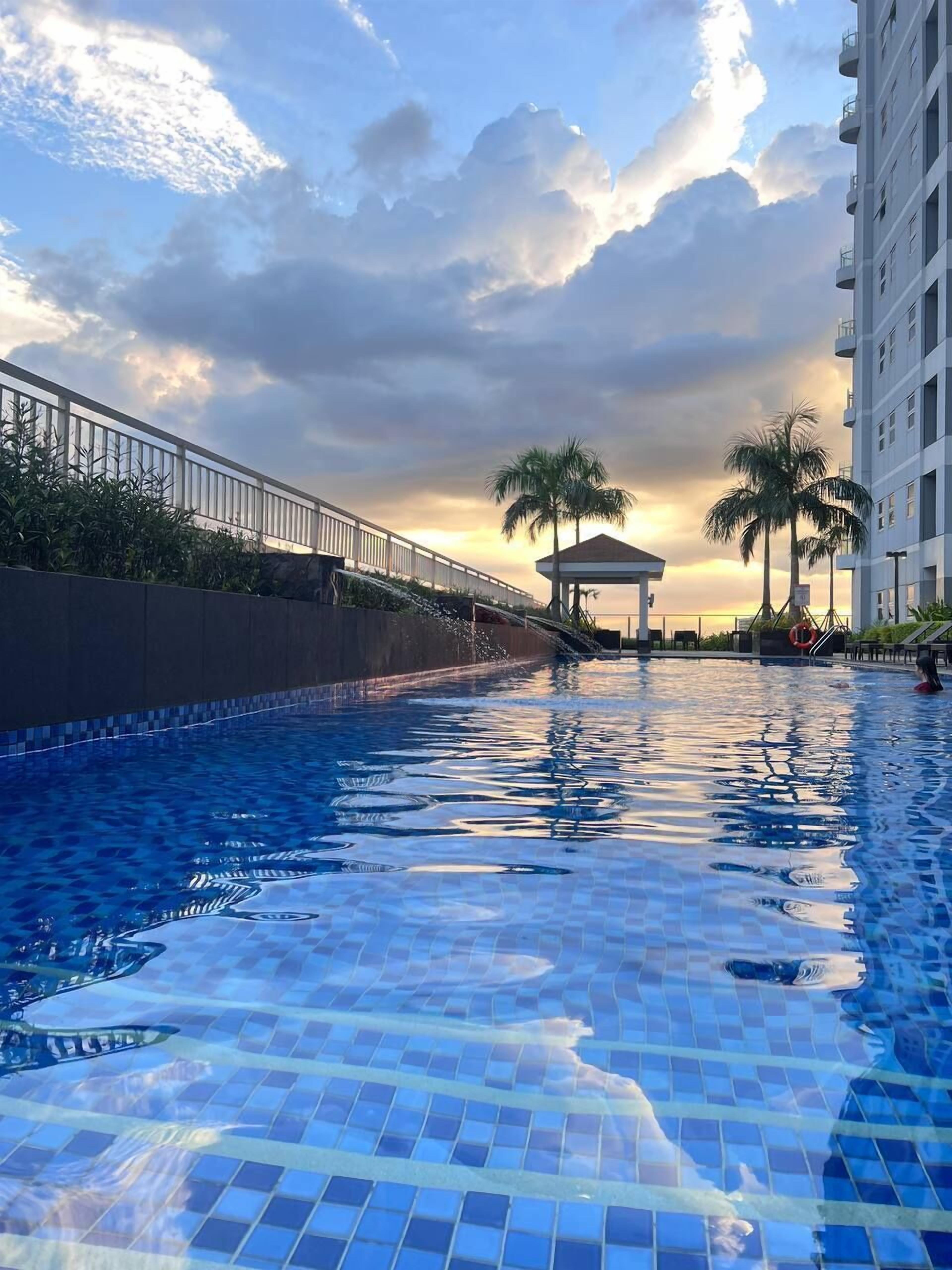 A swimming pool with tiled flooring, a white fence, palm trees, and a building in the background.