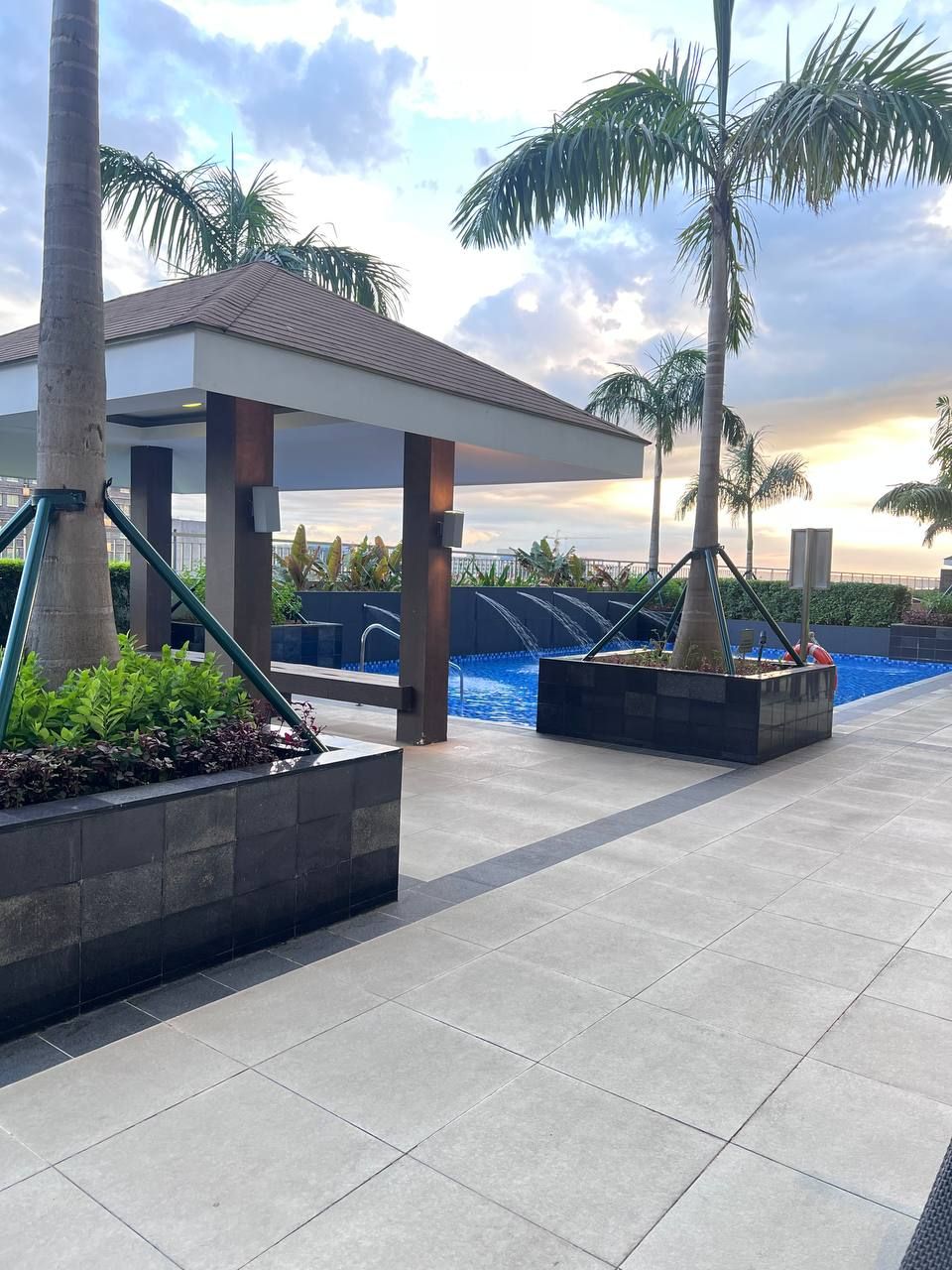 Uma área de piscina com espaço de estar coberto, palmeiras e um pôr do sol ao fundo.
