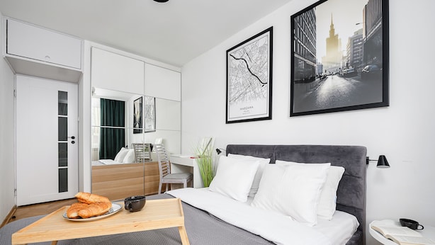 A modern hotel room with a bed, bedside table, and a desk with a chair. There are framed pictures on the wall and a window with a view of a cityscape. - Lumina 2-room apart near Złote Tarasy (Warsaw)