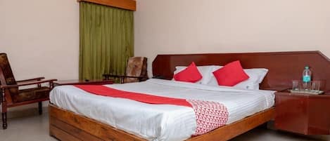 Una habitación de hotel con una cama de madera, almohadas rojas y blancas, una cortina verde, una silla de madera y una mesita de noche con vasos y una botella.