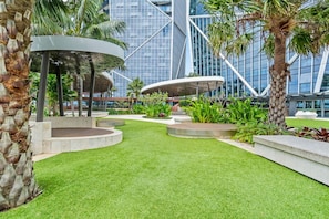 A landscaped outdoor area with a circular seating area, palm trees, and a modern building with glass facade in the background.