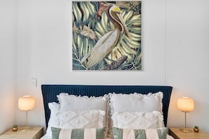 A bed with a blue headboard, two bedside lamps, and a framed artwork above it.