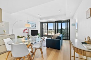 A modern living room with a glass dining table, white chairs, a blue sofa, and a large window offering a city view.