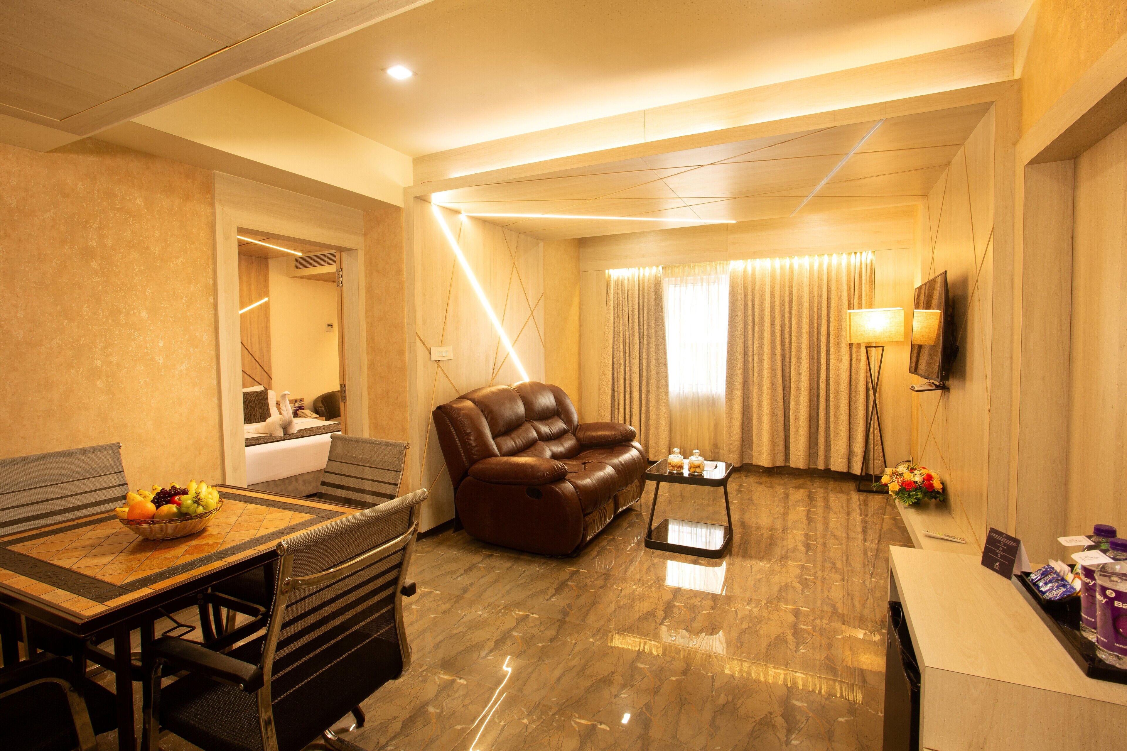 A hotel room with a dining table, a leather sofa, a small coffee table, a TV, and a bathroom with a mirror and sink.