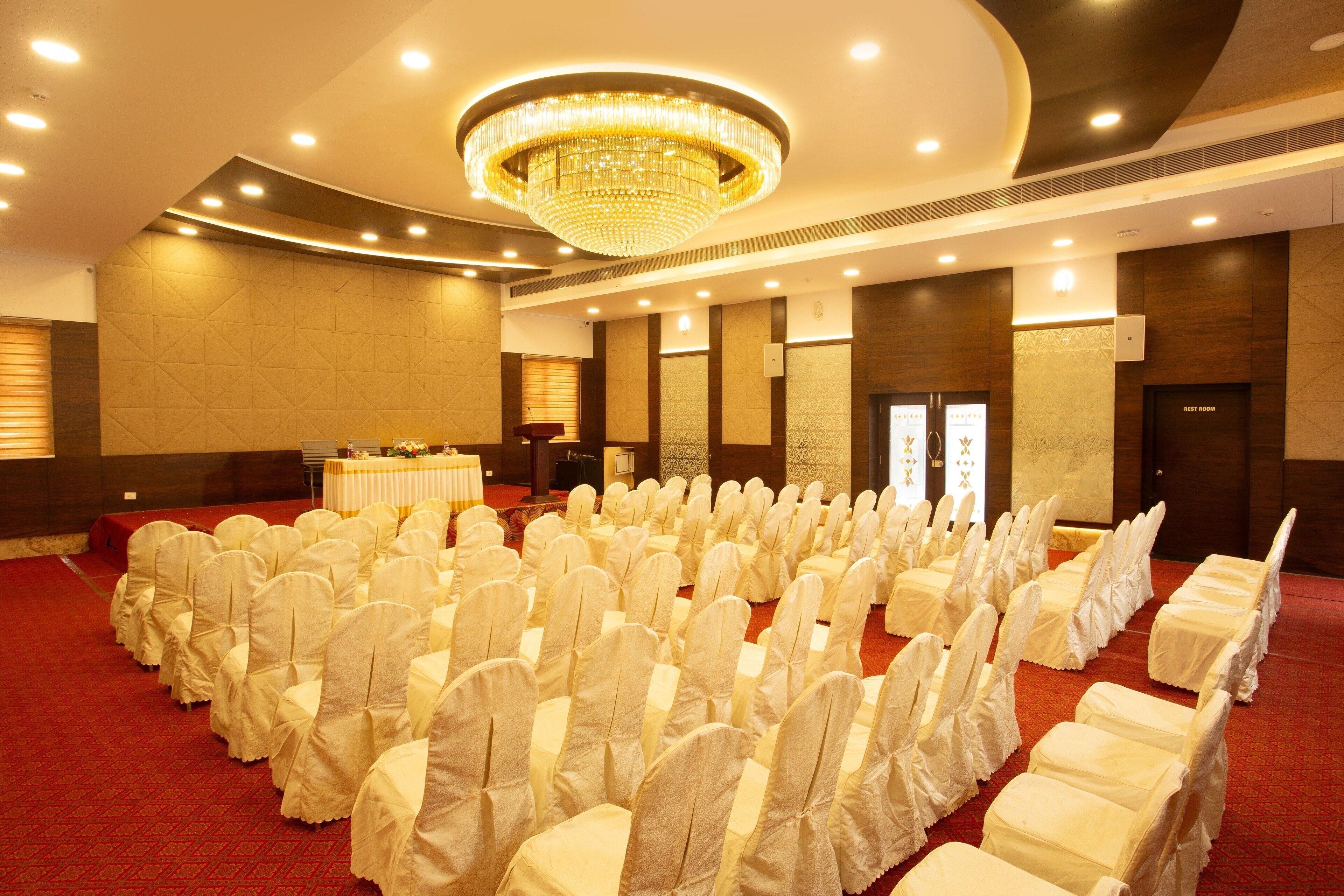 A banquet hall with rows of white-covered chairs, a central stage with a table, and a large chandelier.