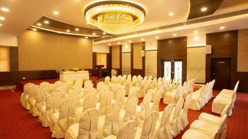 A banquet hall with rows of white-covered chairs, a central stage with a table, and a large chandelier.