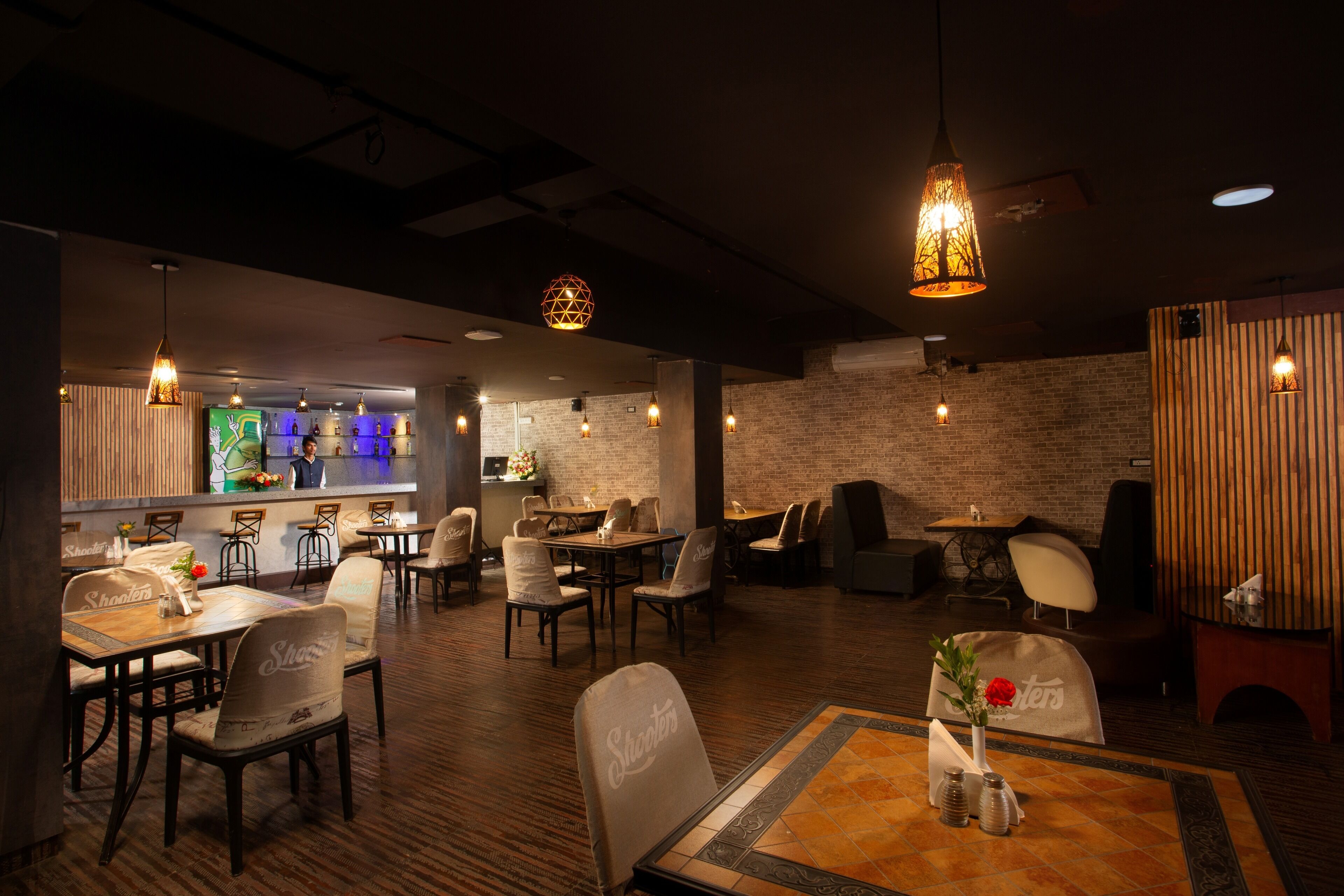 A restaurant with wooden tables and chairs, hanging pendant lights, and a brick wall.