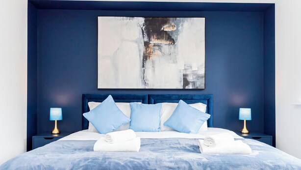 Una cama bien hecha con ropa de cama azul, dos lámparas de noche y un cuadro abstracto grande en la pared.