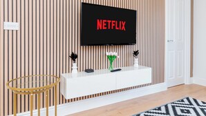 Una sala de estar moderna con un televisor de pantalla montado que muestra 'NETFLIX', una consola blanca con objetos decorativos, una mesa redonda de vidrio y una alfombra estampada.