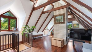 Amplia habitación en el ático con vigas de madera, chimenea y un ventanal con vistas a la vegetación.