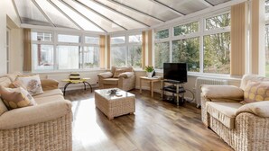 A sunlit living room with wicker furniture, a television, and large windows offering a view of greenery.