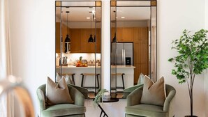 A modern living room with two green armchairs, a small coffee table, and a potted plant. A large mirror reflects a kitchen with wooden cabinets and a refrigerator. - Marbella Lane Nohea Stylish Home Makaha (Waianae)