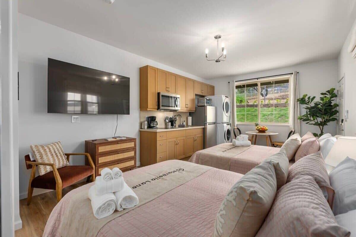 A hotel room with two beds, a TV, a kitchenette, and a window with a view of greenery.