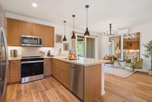 Una cocina moderna con armarios de madera, electrodomésticos de acero inoxidable, iluminación colgante y vistas a un comedor.