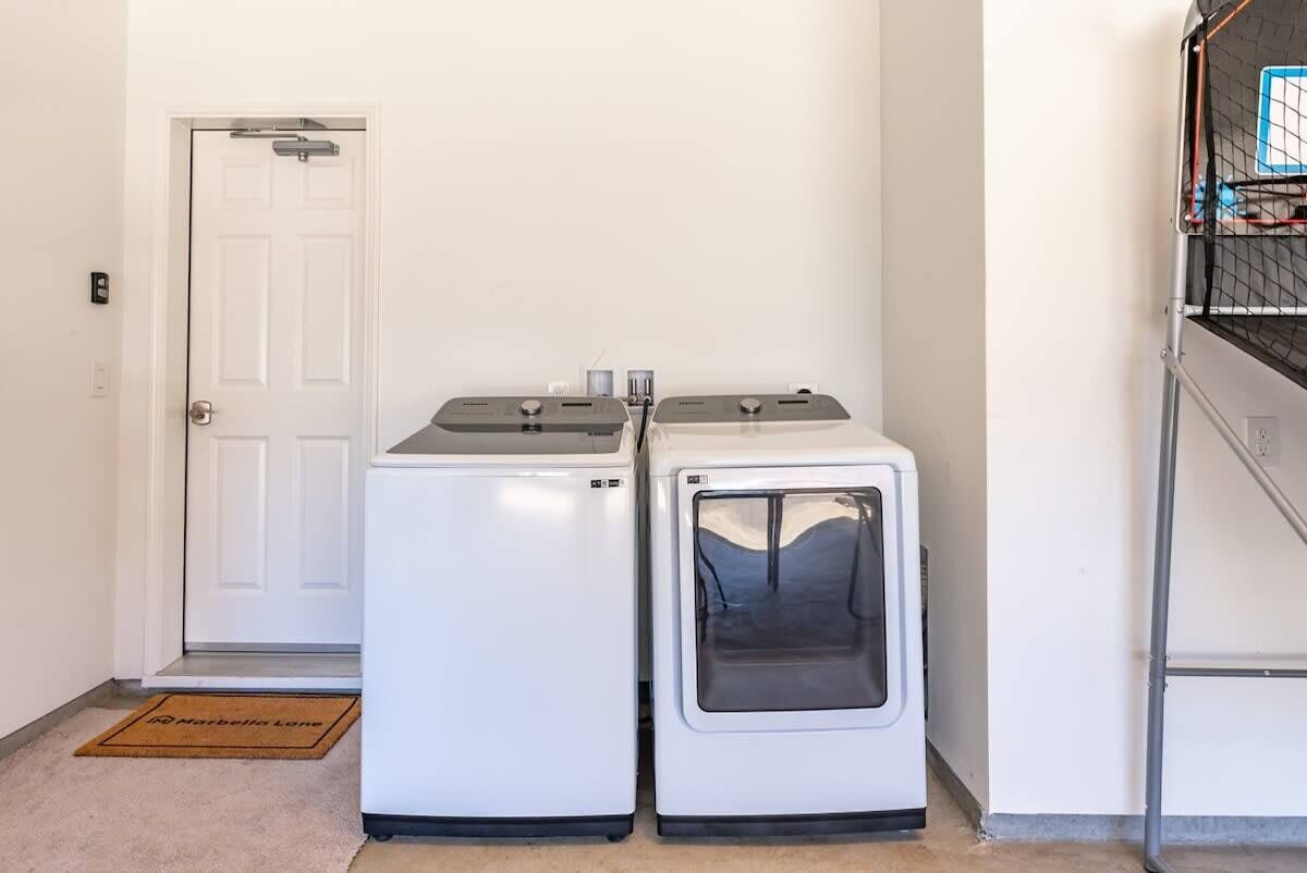 Two white front-loading washing machines in a hallway with a white door and a metal rack with items on the right.