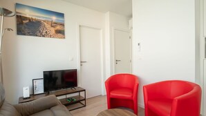 Un salon moderne avec un canapé, deux fauteuils rouges, une table basse, un téléviseur posé sur un meuble et un tableau accroché au mur.