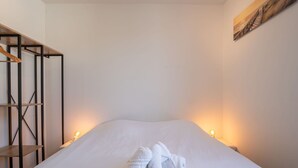 Un lit bien fait, avec deux serviettes, une étagère en bois et un tableau accroché au mur.