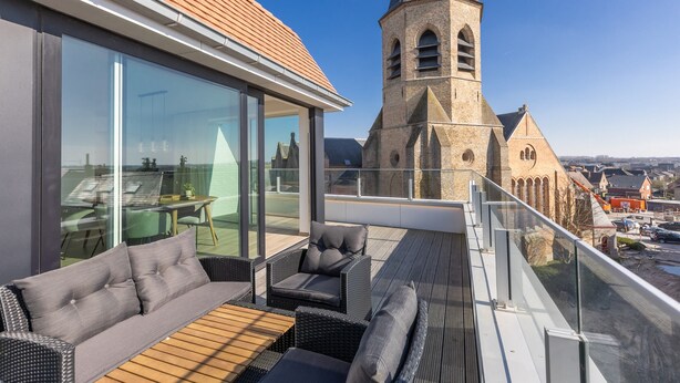 Un balcón moderno con sofá, sillas y una mesa, con vistas a una torre de iglesia histórica y al paisaje urbano.