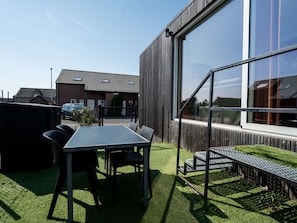 Ein modernes Gebäude mit großen Fenstern, einer Terrasse mit Tisch und Stühlen sowie ein geparktes Auto im Hintergrund.