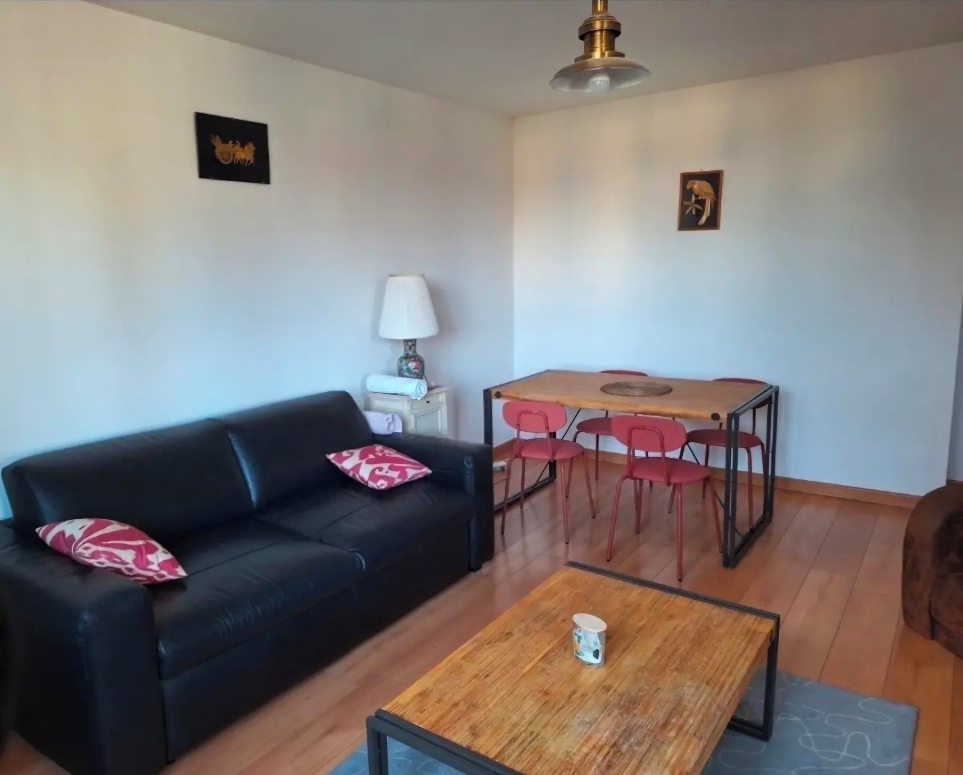Een woonkamer met een zwarte bank, een houten salontafel, een eettafel met rode stoelen, een lamp en twee aan de muur bevestigde foto's.