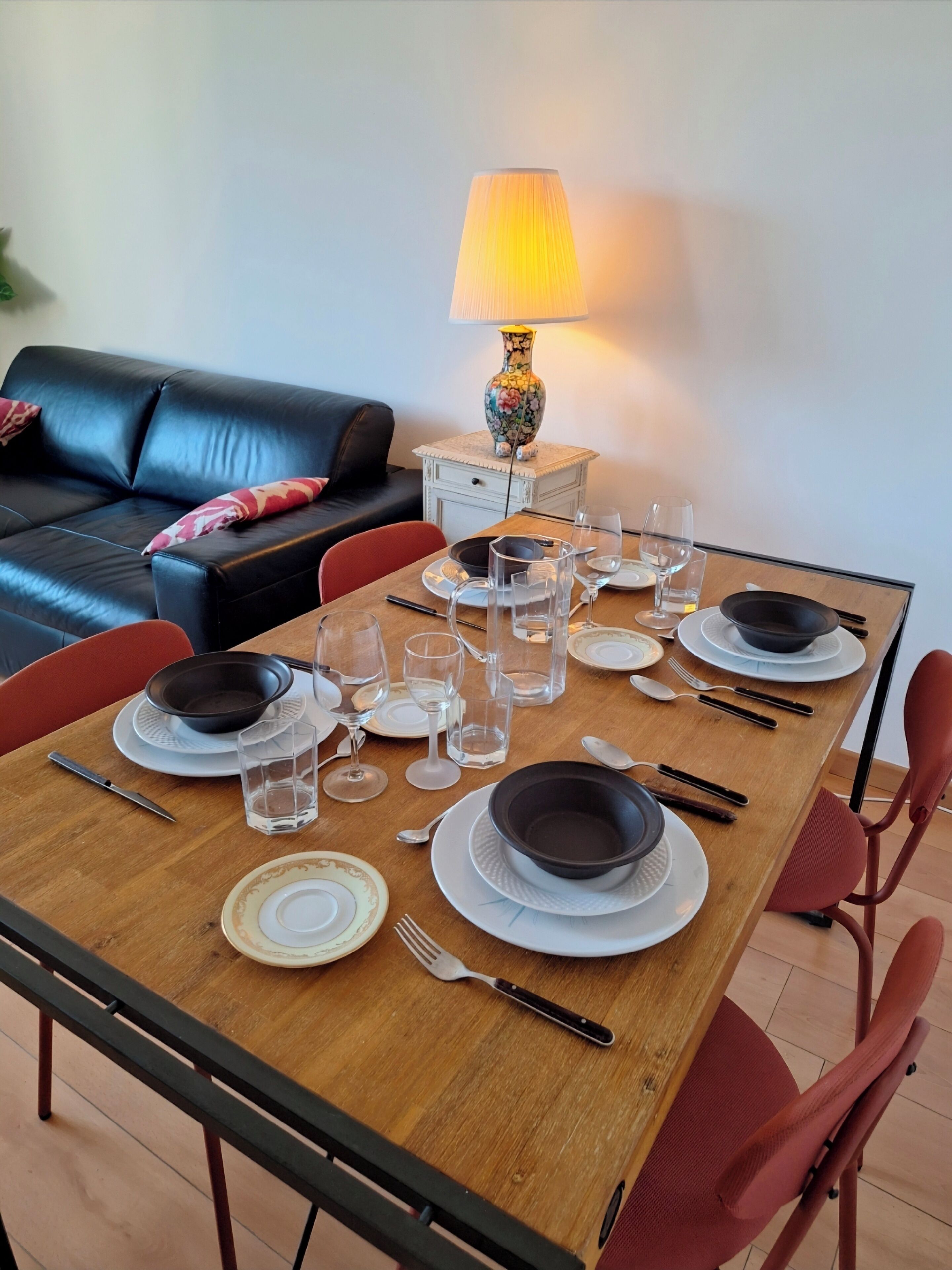 A dining table set with plates, bowls, glasses, and cutlery, with a sofa and a lamp in the background.
