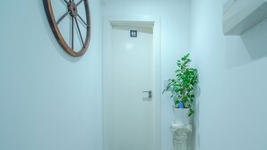 Un couloir étroit orné d’une roue en bois, une porte avec une indication pour les toilettes hommes et femmes, une plante en pot et une colonne blanche.