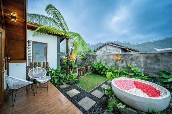A modern outdoor patio with a wooden deck, two white wicker chairs, a concrete planter with red and white pebbles, and lush greenery.