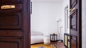 Un couloir avec une porte en bois sombre, un banc recouvert d’un tissu blanc, une petite table ronde avec une lampe et un tapis à motifs.