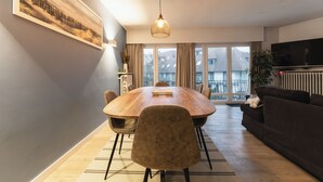 Ett modernt vardagsrum med ett matbord i trä, bekväma sittplatser, ett stort fönster med gardiner och en tv med platt skärm.