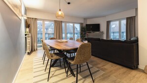 Ett modernt vardagsrum med ett matbord i trä, bekväma sittplatser, ett stort fönster med gardiner, en svart soffa och en tv.