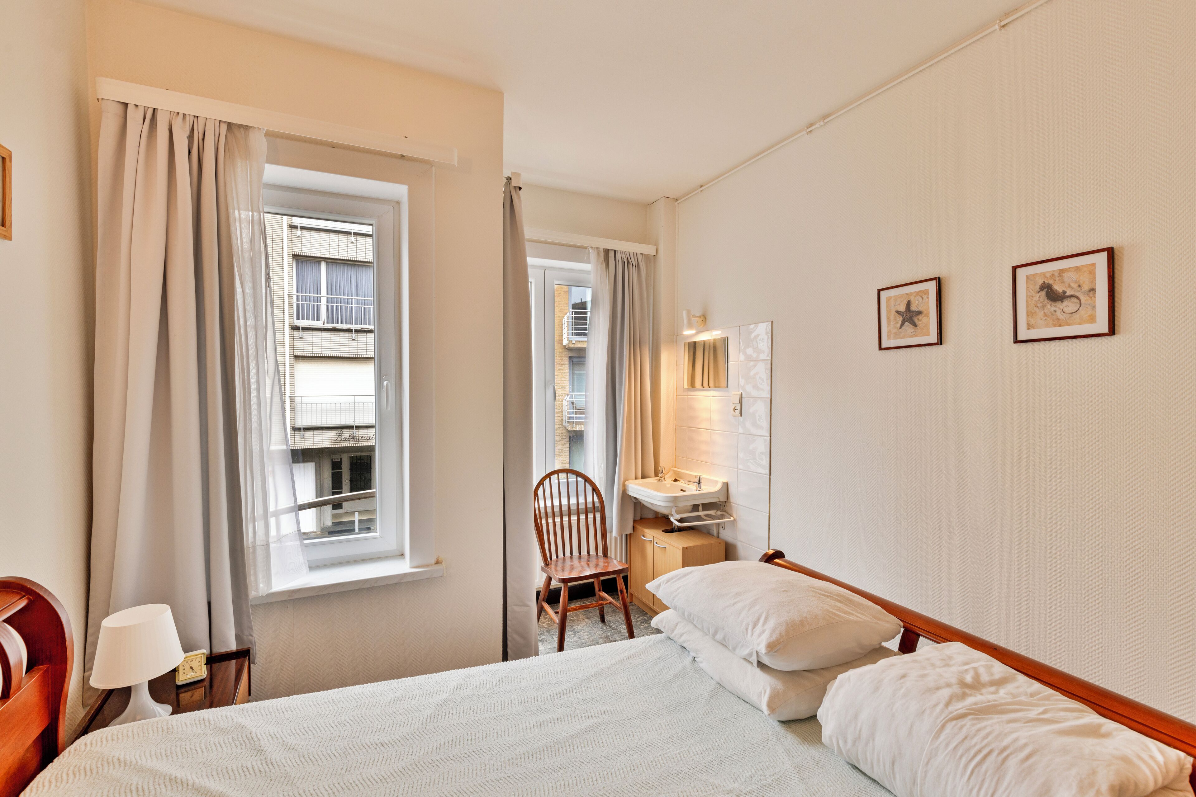 A hotel room with a bed, a chair, a desk with a lamp, a window with curtains, and framed pictures on the wall.