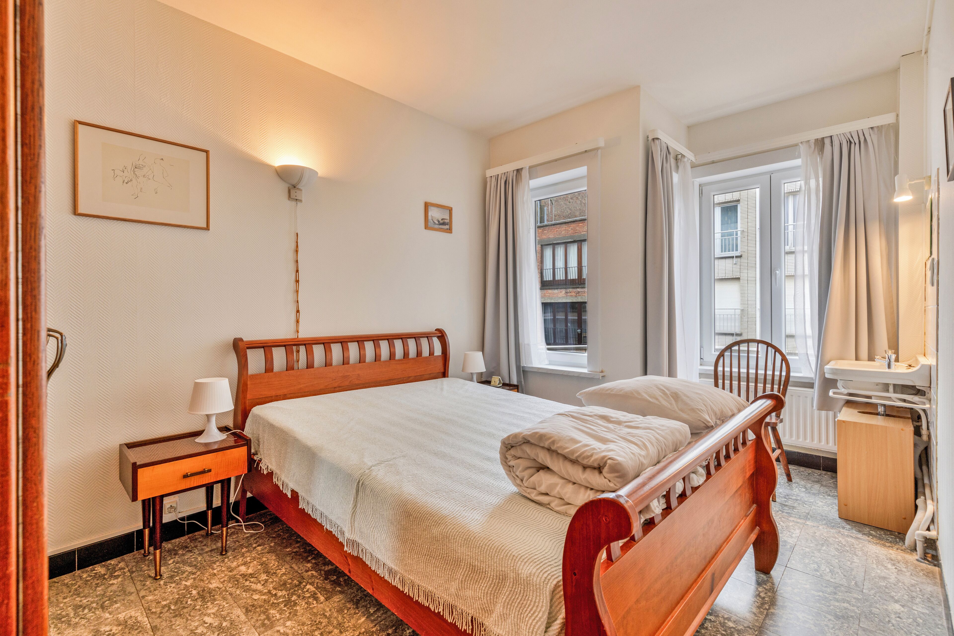 Un dormitorio ordenado con una cama de madera, una mesita de noche con lámpara, una pequeña silla y una ventana con cortinas.