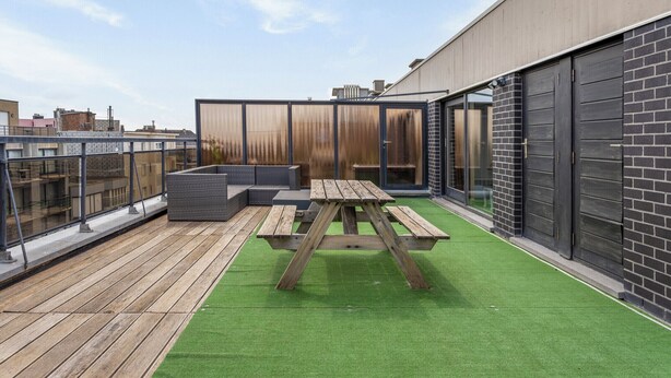 Terraza en la azotea con mesa de picnic de madera y zona de estar, rodeada por una barandilla de cristal y vistas a la ciudad.
