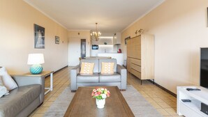 Een moderne woonkamer met een grijze bank, een bijzettafeltje met een bloevas, een televisie op een dressoir en een keuken in de achtergrond.