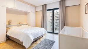 Een slaapkamer met een bed, nachtkastje, dressoir en uitzicht op een balkon.