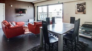 A modern living room with a red sofa, a dining table with chairs, a TV mounted on the wall, and a view of the outside through large windows. - Gouden Boom 0618 + P* (Koksijde)