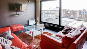 A modern living room with red leather sofas, a glass coffee table, a flat-screen TV, and a large window offering a city view. - Gouden Boom 0618 + P* (Koksijde)